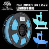A spool of PLA luminous 1KG 1.75mm filament in luminous blue, shown alongside two small 3D-printed boat models demonstrating the glow effect, on a black background with the SpoolSpider brand logo.