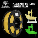 SpoolSpider PLA Luminous Yellow 1KG 1.75mm filament on a black spool, shown alongside two small 3D-printed toy boat models in glowing yellow and glowing green on a black background.